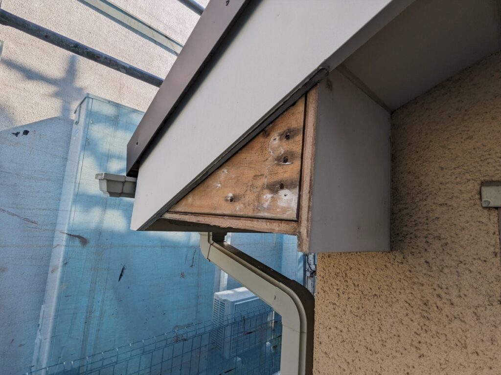 Close-up of a weathered wooden beam under a gray metal overhang above a beige stucco wall, with a downspout and blue building in the background.