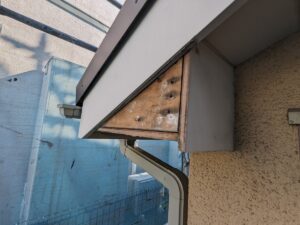 Close-up of a weathered wooden beam under a gray metal overhang above a beige stucco wall, with a downspout and blue building in the background.