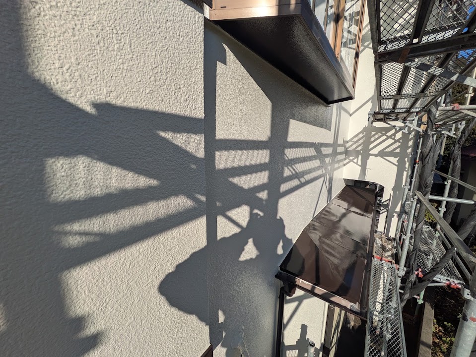 07-wall-detail | 外壁塗装は神奈川県横浜市の塗装職人 Shadowy scaffolding cast long geometric shadows on a white exterior wall beside a construction site; metal platforms and rails are visible above an opening of the building. 10:30 AM sunlight.