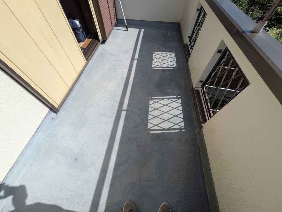 10-balcony-before | 外壁塗装は神奈川県横浜市の塗装職人 Narrow outdoor balcony with a concrete floor, beige walls, and metal railing casting geometric shadows on the floor.