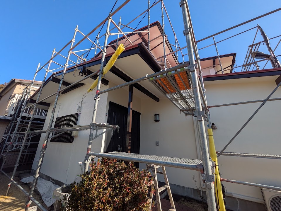 12-exterior-detail | 外壁塗装は神奈川県横浜市の塗装職人 House under exterior renovation with metal scaffolding surrounding the facade, blue sky above, and a small bush in front.