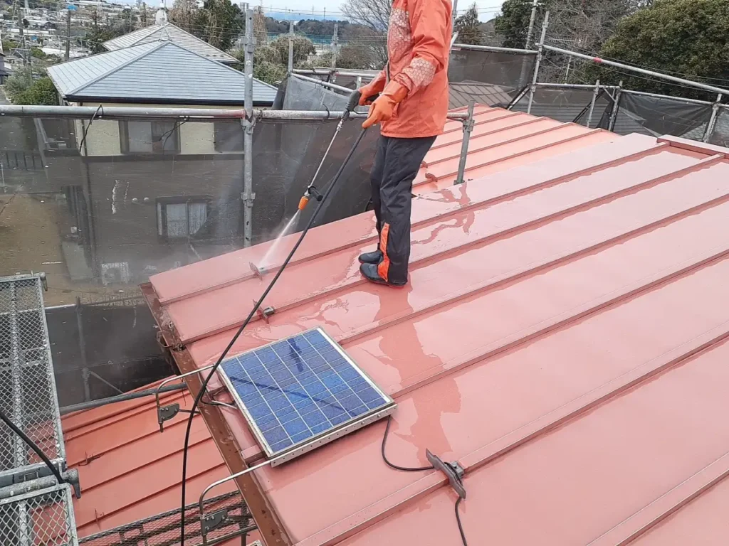 13-roof-washing | 外壁塗装は神奈川県横浜市の塗装職人 Worker in orange gear pressure-washing a red metal roof on scaffolding