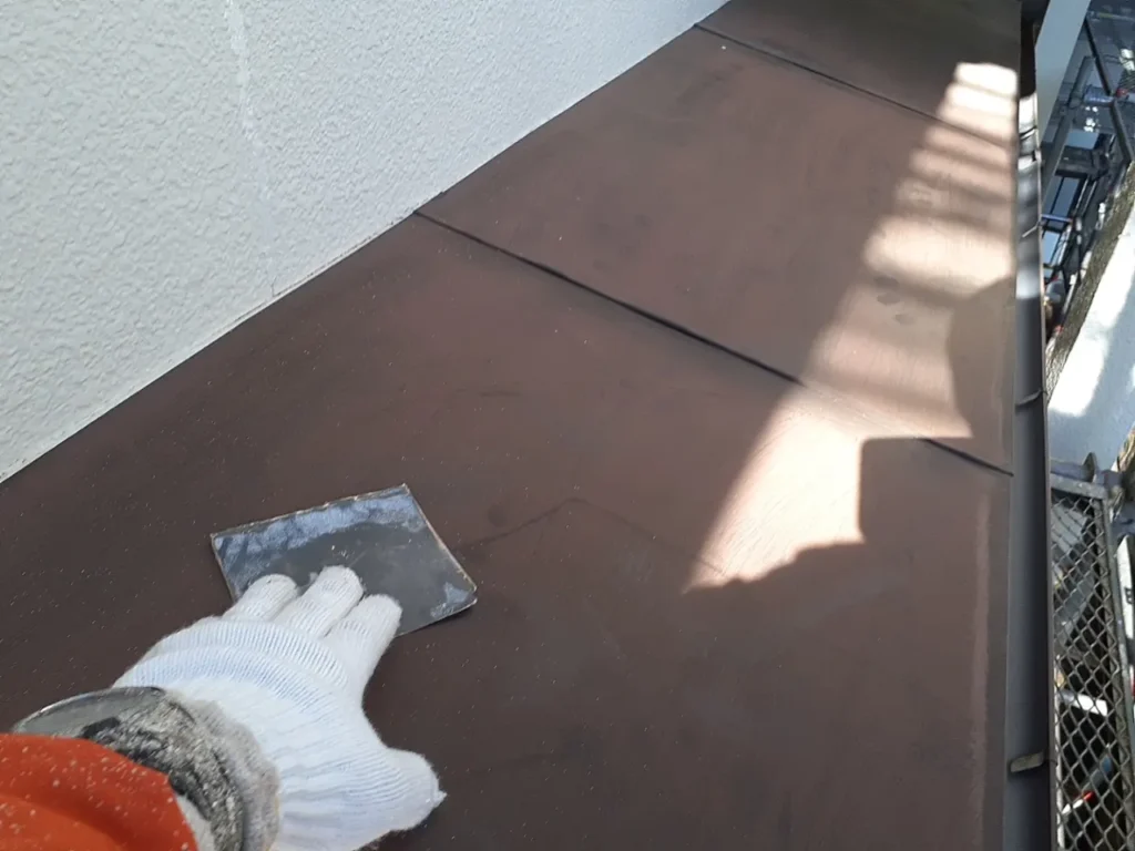 14-eave-preparation | 外壁塗装は神奈川県横浜市の塗装職人 Gloved hand with a scraper smoothing a brown metal roof panel near a white wall and scaffolding.