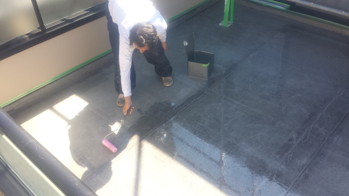 METADATA-START | 外壁塗装は神奈川県横浜市の塗装職人 Worker applying waterproof coating to a flat rooftop using a pink paint roller, with a bucket nearby under bright sunlight.