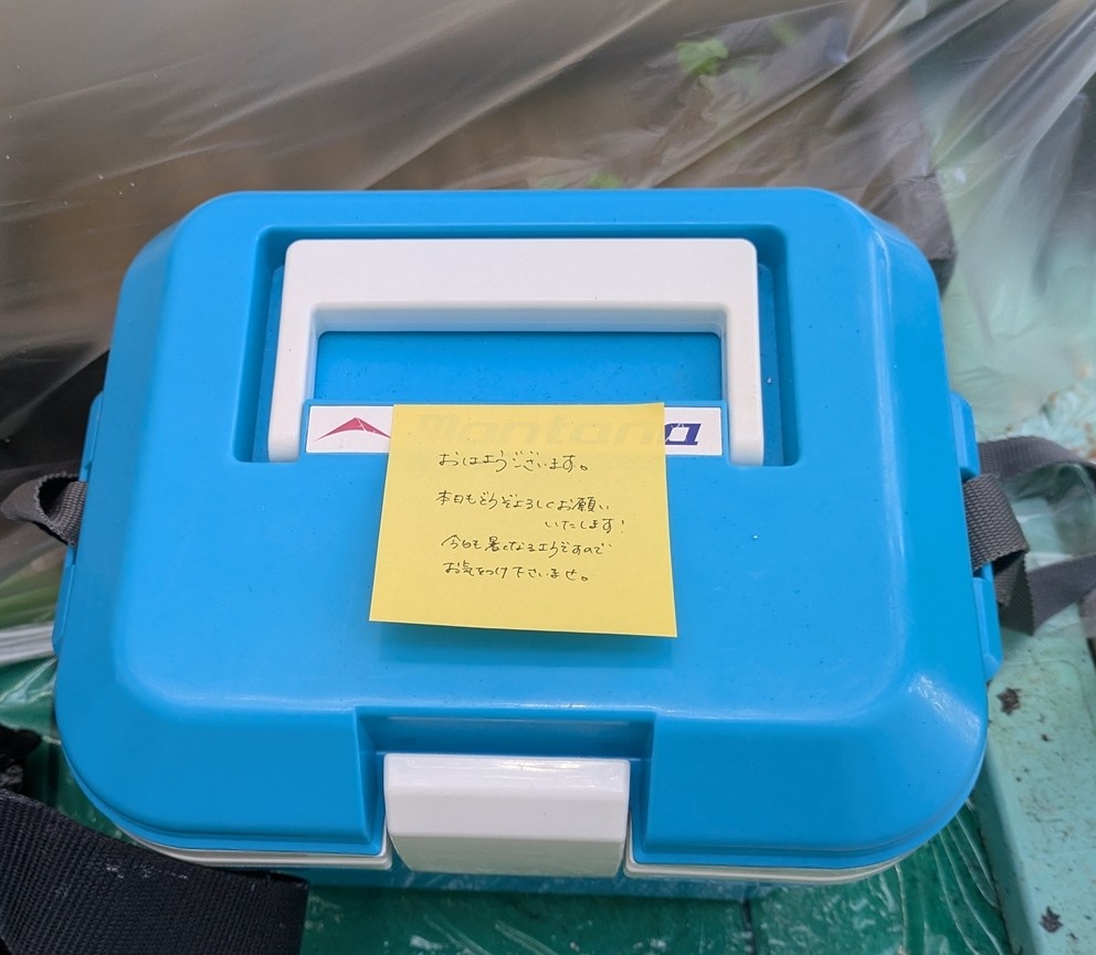 PXL_20260519_024240086 | 外壁塗装は神奈川県横浜市の塗装職人 Blue plastic toolbox with a white inner lid and a yellow sticky note attached to the top edge, resting on a green surface with a strap visible on the side.