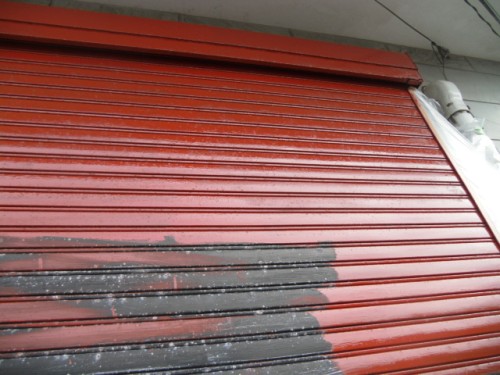 koushin_CIMG1291-1 | 外壁塗装は神奈川県横浜市の塗装職人 Closed red corrugated rolling shutter door, likely at a storefront, with a dark gray patch near the bottom and a white exterior wall to the right.