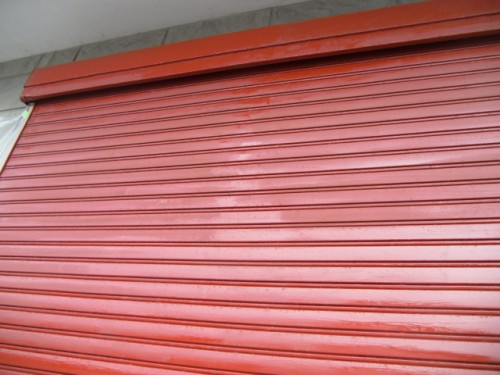 koushin_CIMG1293-1 | 外壁塗装は神奈川県横浜市の塗装職人 Closed red metal rolling shutter covering a storefront or garage door, with horizontal slats and a small overhang at the top.