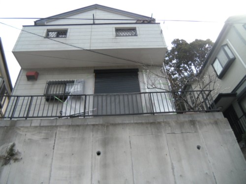 koushin_CIMG1303-1 | 外壁塗装は神奈川県横浜市の塗装職人 Two-story white house perched over a concrete retaining wall with a metal railing; small barred windows on the upper level and a large roll-up door on the lower level, with a tree visible on the right.