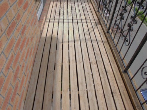 Wooden slatted balcony deck running along a brick wall with decorative iron railing on the right.
