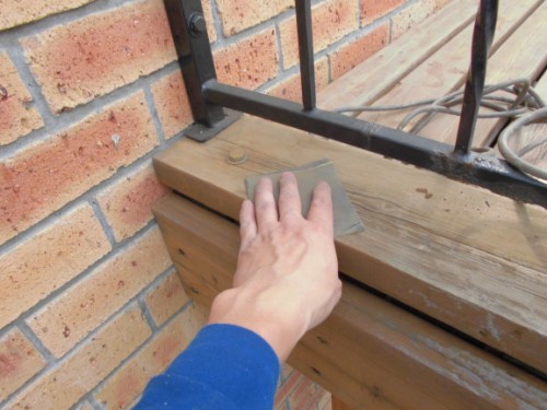 Person in a blue sleeve smoothing wood filler along a wooden railing with a putty knife next to a brick wall.