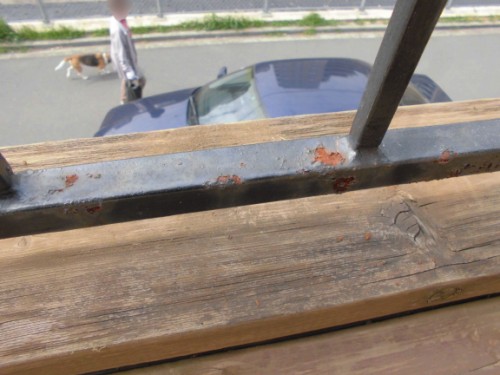 View from a weathered wooden balcony railing: a blue car on the street below, a person walking a small dog in the distance, with rust on the metal strip.
