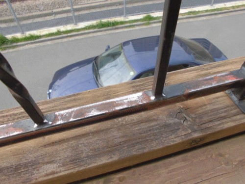 View from a wooden balcony railing with metal bars, looking down on a blue car parked along a street.