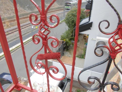 Red wrought-iron railing with decorative scrolls on a balcony, overlooking a street and railway tracks below.