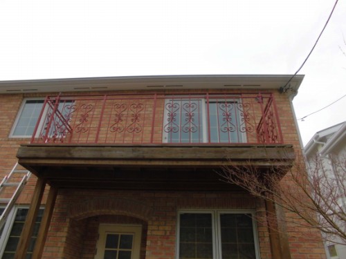 Brick two-story house with a second-floor balcony featuring red decorative iron railing above the entryway.