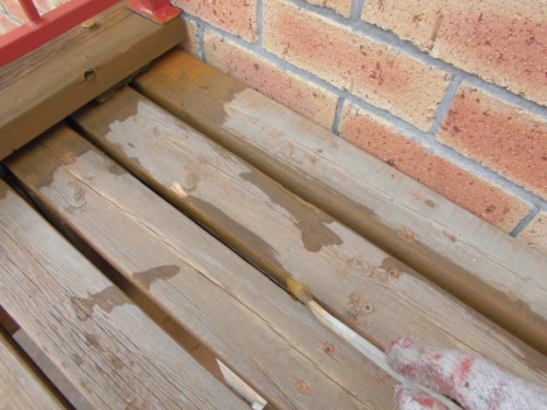 Wet wooden outdoor boards beside a brick wall, with a gloved hand wiping one plank.