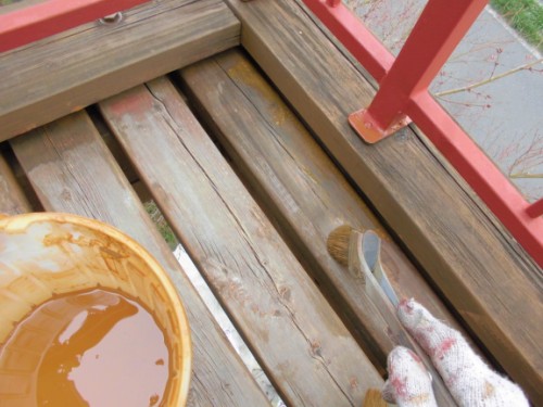Gloved hand holding a brush over a wooden deck railing beside a bucket of brown wood stain.