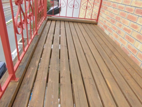 Outdoor wooden balcony/deck with parallel planks, a red decorative metal railing on the left, and a brick wall on the right; boards show weathering and gaps between them.