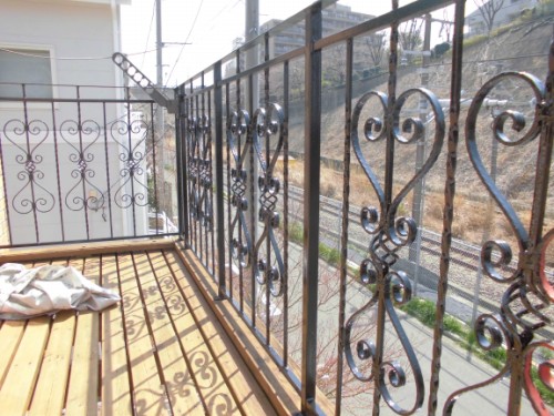 Balcony with decorative wrought-iron railing and wooden planks; a folded cloth on the left, with train tracks and a hillside beyond.