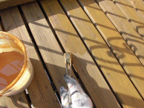 Gloved hand holding a scraper over wooden deck boards, with an orange paint bucket nearby.