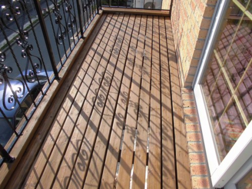 Wooden balcony deck with vertical boards, ornate black railing, brick wall, and a glass door on the right.