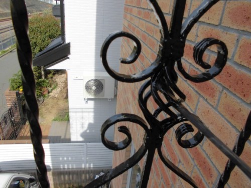 Close-up of an ornate black wrought-iron railing on a brick balcony, with a white air conditioner unit mounted on a nearby building in the background and a street below