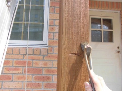 Close-up of a hand holding a hammer and driving a nail into a wooden porch post beside a brick wall and front door.