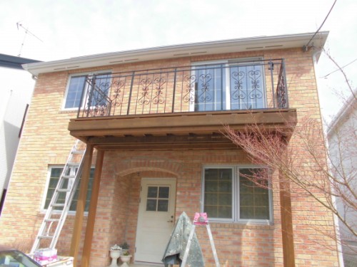 Brick two-story house under renovation with a covered front porch, decorative iron balcony railing on the second floor, and a ladder leaning on the left.