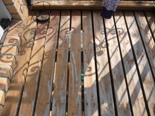 Weathered wooden deck with peeling paint and gaps between slats; ornate railing shadows cast on the boards, brick wall on the left, and a small potted plant visible near the top-left.