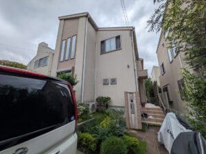 Front view of a two-story beige house with tall narrow windows, a small garden, and a white van in the foreground.