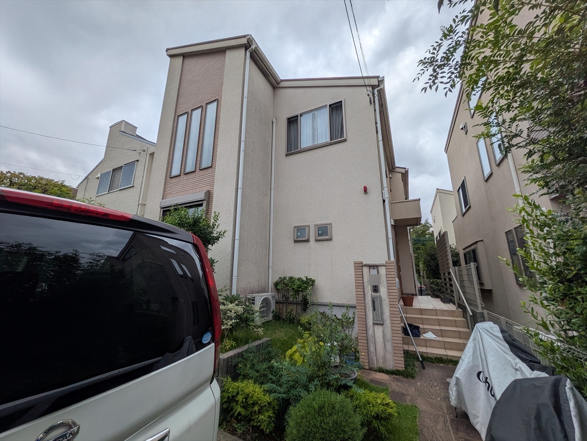 Front view of a two-story beige house with tall narrow windows, a small garden, and a white van in the foreground.