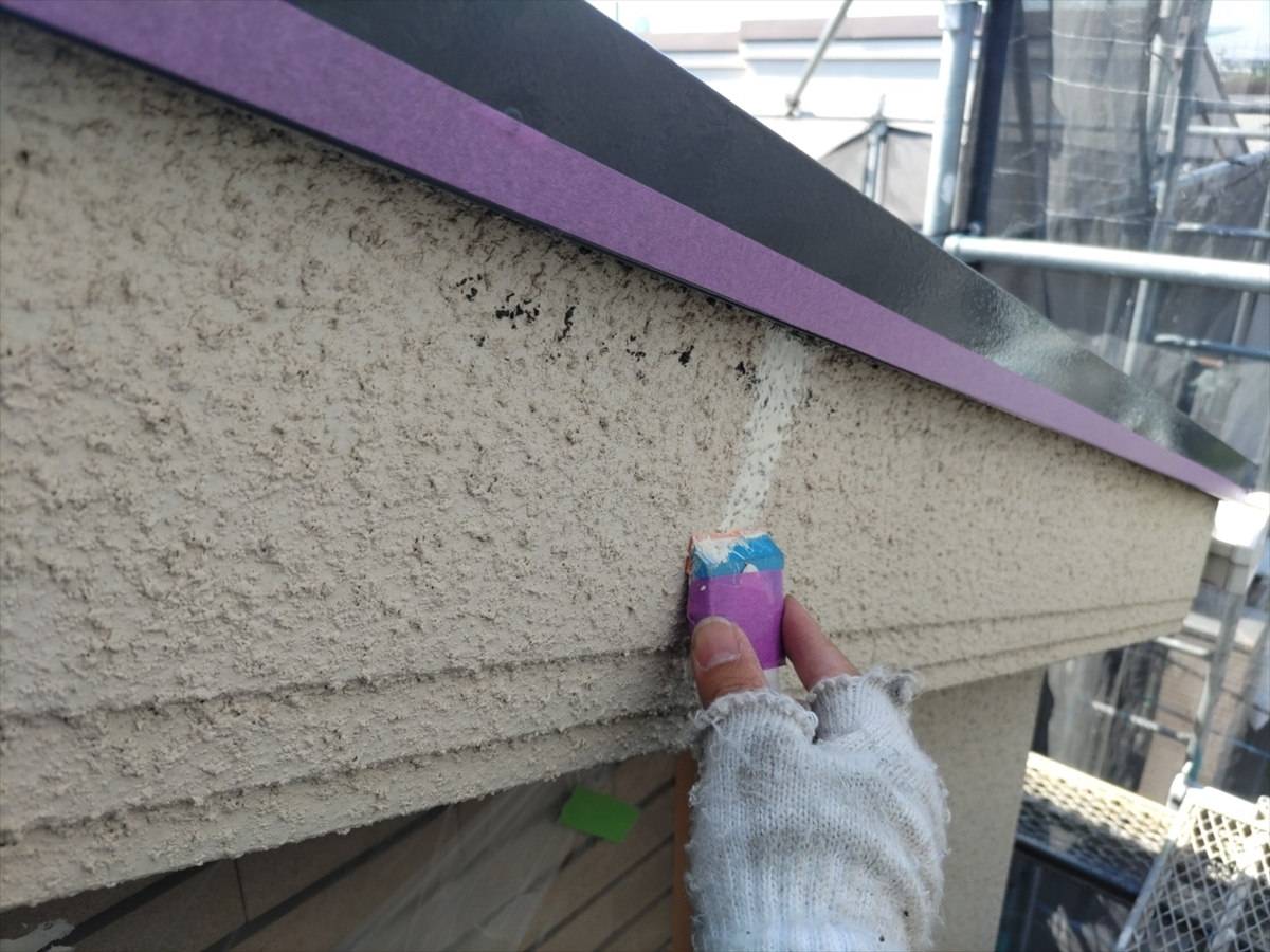 Gloved hand applying white sealant along a beige textured exterior wall, with purple painter’s tape and construction scaffolding in the background.