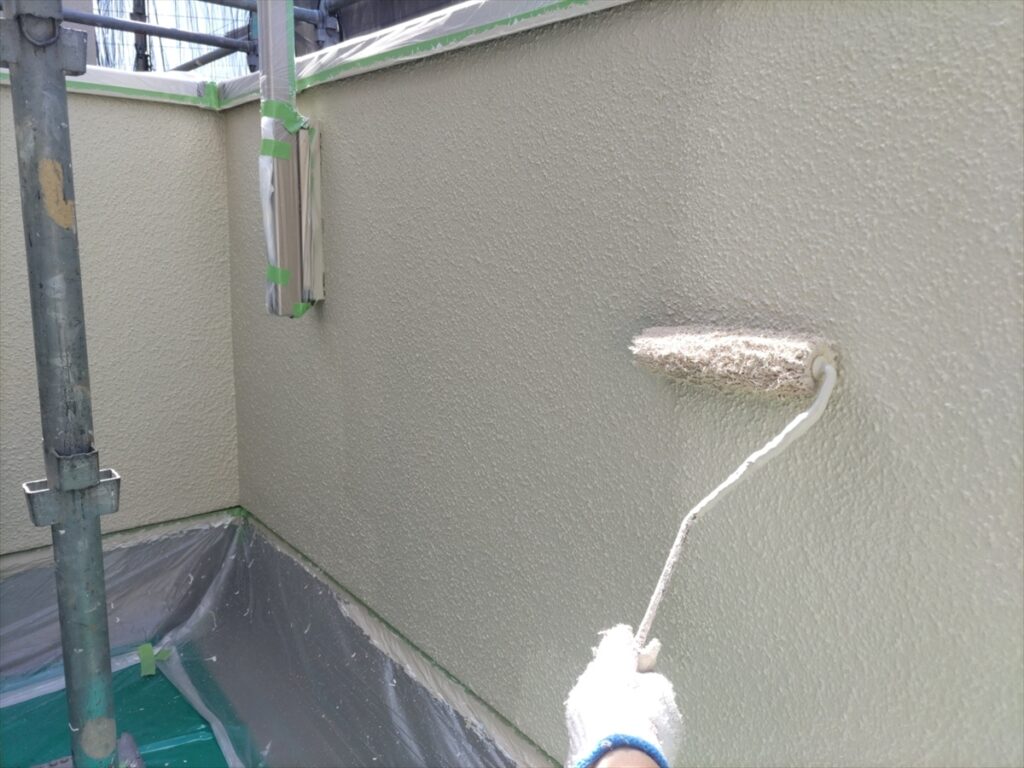 Painter applies beige textured paint to an exterior wall with a roller, scaffolding and drop cloth nearby.