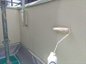 Painter applies beige textured paint to an exterior wall with a roller, scaffolding and drop cloth nearby.