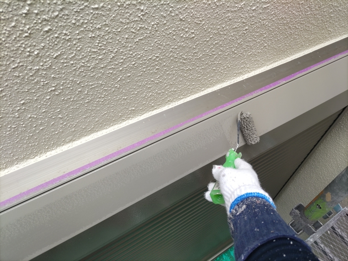 Person wearing gloves painting a metal window/door frame with a small roller; purple painter's tape along the wall edge to protect surfaces.
