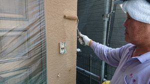 Worker in gloves and head wrap uses a paint roller to apply beige paint to an exterior stucco wall, standing on scaffolding beside a protected door/window.
