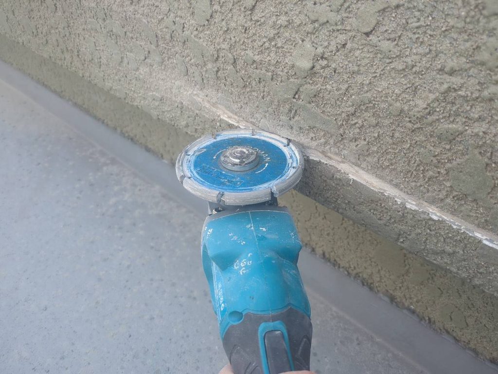写真_260824_39 | 外壁塗装は神奈川県横浜市の塗装職人 Blue handheld angle grinder smoothing a rough concrete wall during construction. A mortar line is visible along the edge of the wall.