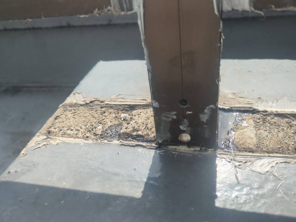 写真_260824_43 | 外壁塗装は神奈川県横浜市の塗装職人 Close-up of a vertical metal post bolted to a concrete rooftop, with peeling paint and sealant around the base.