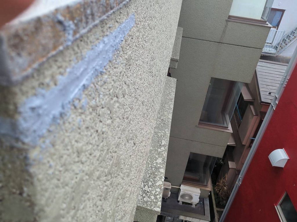 写真_260826_27 | 外壁塗装は神奈川県横浜市の塗装職人 View of a gray apartment building with windows and an outdoor AC unit at ground level, shot from a narrow alley between walls.