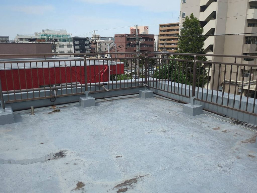 写真_260826_4 | 外壁塗装は神奈川県横浜市の塗装職人 Rooftop terrace with a metal railing and a blue-gray concrete floor, overlooking a cityscape of buildings and trees in the distance.